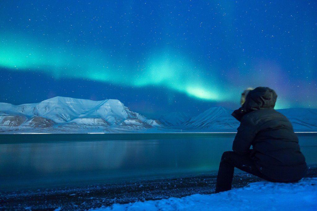A man sits on the shore and looks at the snowy mountains and the Northern Lights above them.