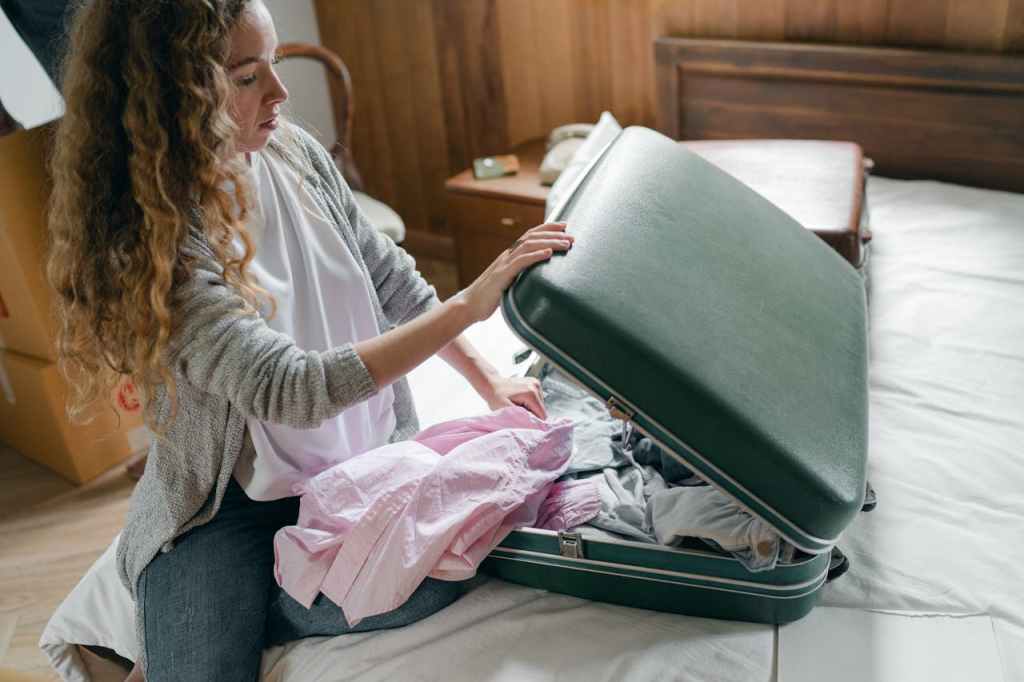 A woman packing a suitcase with clothes and other items on a bed in a cozy room.