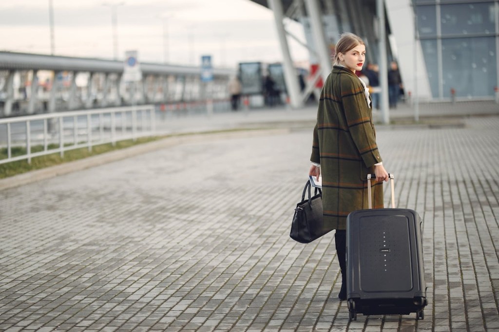 : Woman in a long coat pulling a suitcase, standing outdoors near a modern building, looking back

