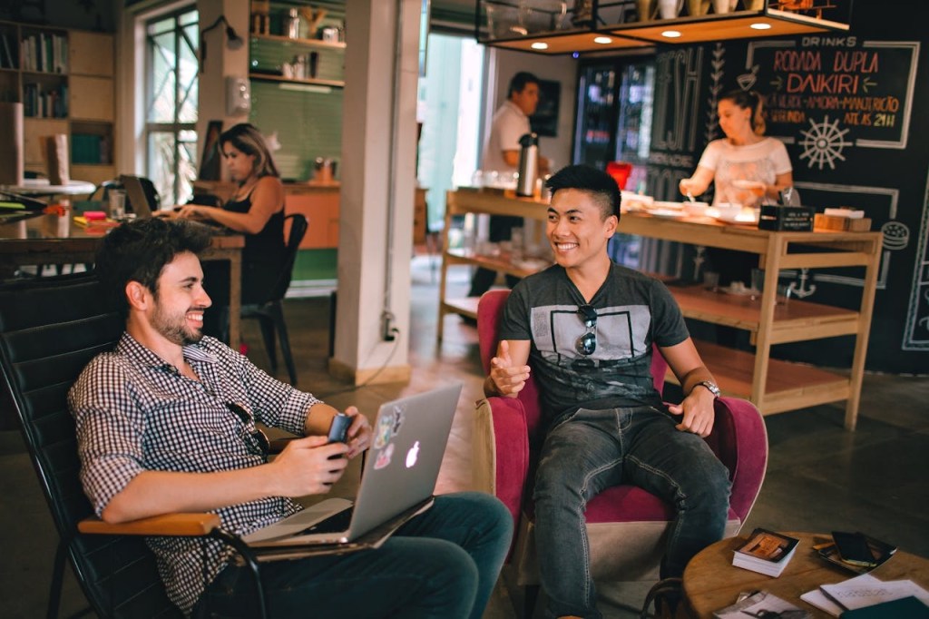 Two young men chatting and smiling while sitting in a cozy café with laptops and phones