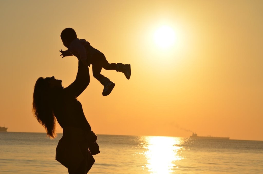 A mother lifting her baby in the air at the beach during sunset, with the sun reflecting on the wate