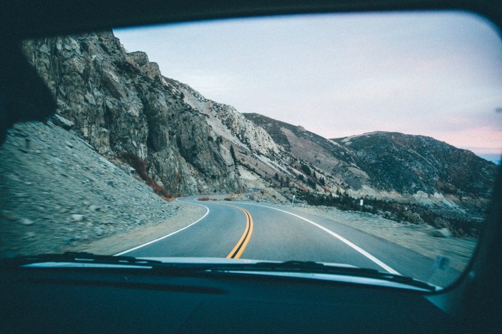 Photograph taken from inside a vehicle showing a winding mountain road with rocky cliffs on the left and a guardrail on the right