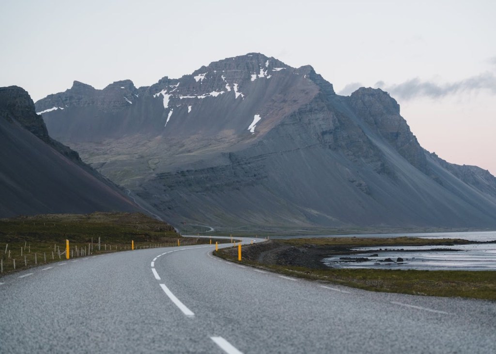 winding road along a coastline with large, rugged mountains in the background, partially covered with snow.
